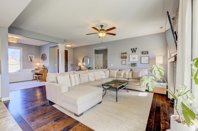 living area featuring hardwood / wood-style floors, ornate columns, wainscoting, a decorative wall, and a ceiling fan