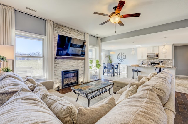 living room featuring baseboards, visible vents, dark wood-style flooring, ceiling fan, and a stone fireplace