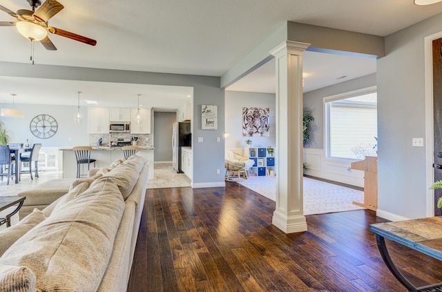 living area with wainscoting, dark wood-style flooring, a ceiling fan, and ornate columns