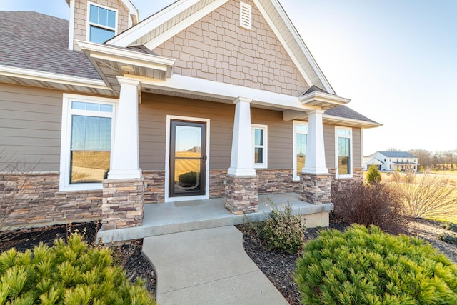 doorway to property with covered porch, stone siding, and roof with shingles