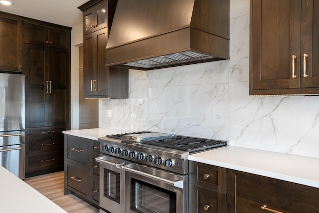kitchen featuring decorative backsplash, dark brown cabinets, custom range hood, light wood-style flooring, and appliances with stainless steel finishes