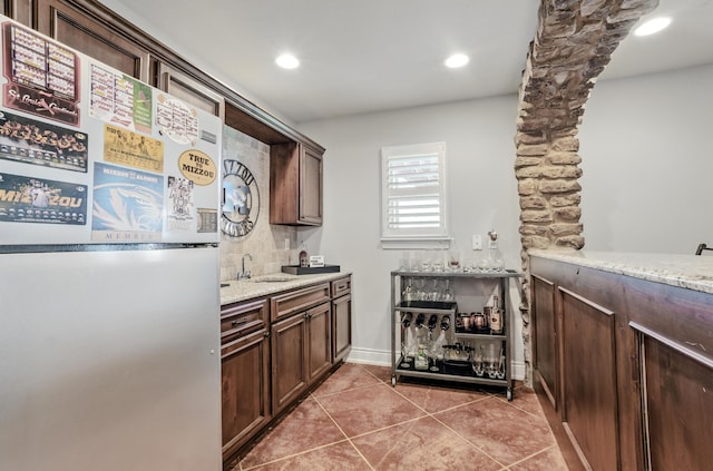 kitchen featuring freestanding refrigerator, light stone countertops, dark brown cabinets, light tile patterned flooring, and a sink