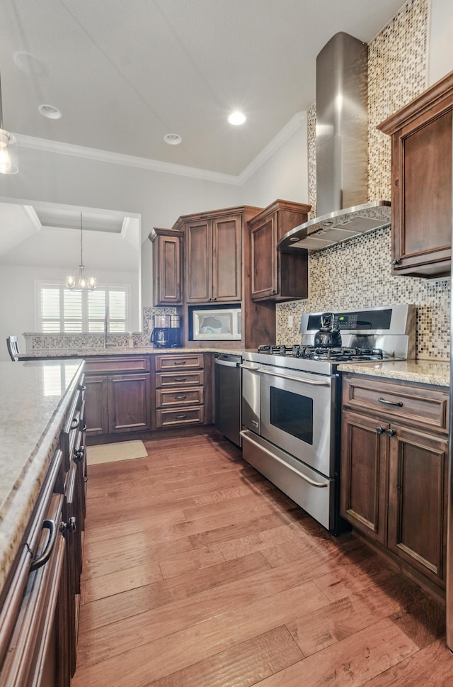 kitchen with a notable chandelier, wall chimney range hood, decorative backsplash, light wood finished floors, and appliances with stainless steel finishes