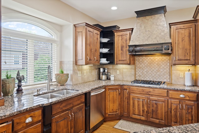 kitchen with light stone countertops, stainless steel appliances, open shelves, custom range hood, and a sink