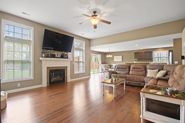 living room featuring wood finished floors, a tile fireplace, ceiling fan with notable chandelier, baseboards, and visible vents