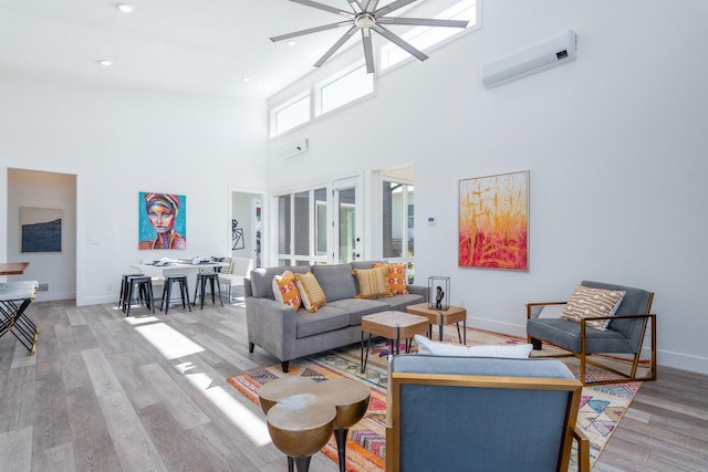 living area with a wall mounted AC, ceiling fan, a towering ceiling, light wood-style flooring, and baseboards