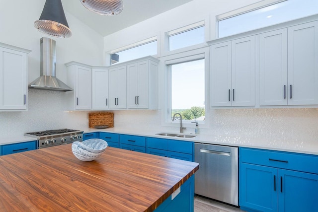 kitchen with appliances with stainless steel finishes, a sink, butcher block counters, blue cabinets, and wall chimney exhaust hood