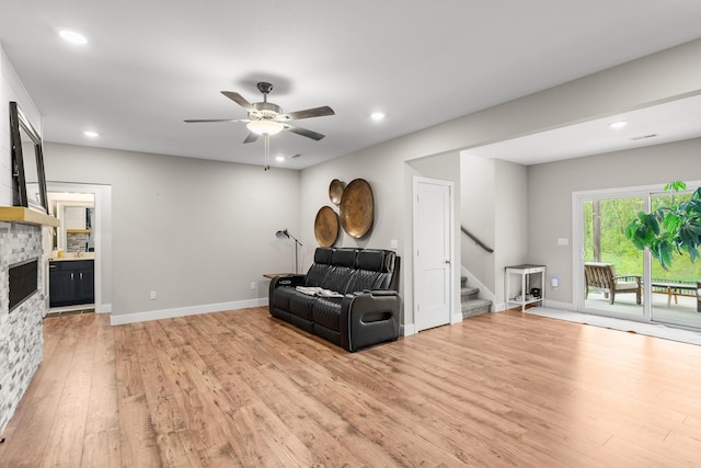 living area featuring a ceiling fan, a stone fireplace, baseboards, light wood-style floors, and stairs
