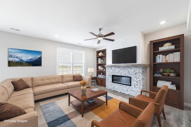 living room with a stone fireplace, light wood-style flooring, ceiling fan, and built in shelves