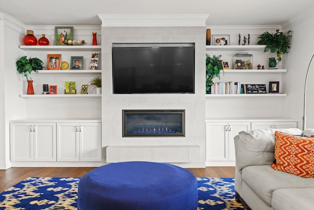living area with wood finished floors, a tiled fireplace, crown molding, and built in shelves