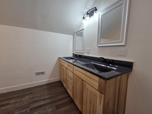 bathroom featuring vaulted ceiling, wood finished floors, double vanity, baseboards, and a textured wall