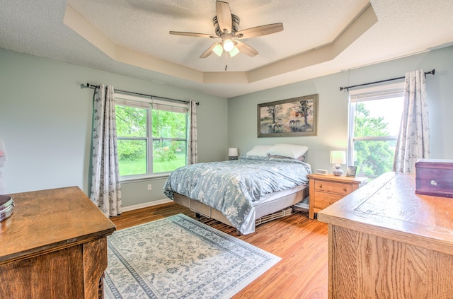 bedroom with a raised ceiling, multiple windows, light wood-style flooring, a textured ceiling, and ceiling fan