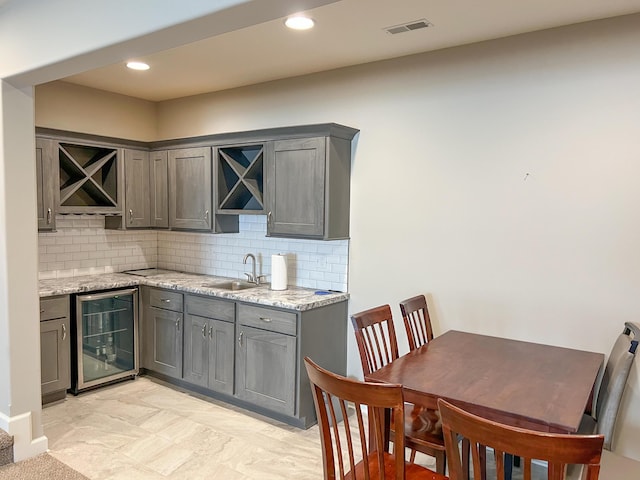 kitchen featuring beverage cooler, gray cabinetry, a sink, backsplash, and light stone counters