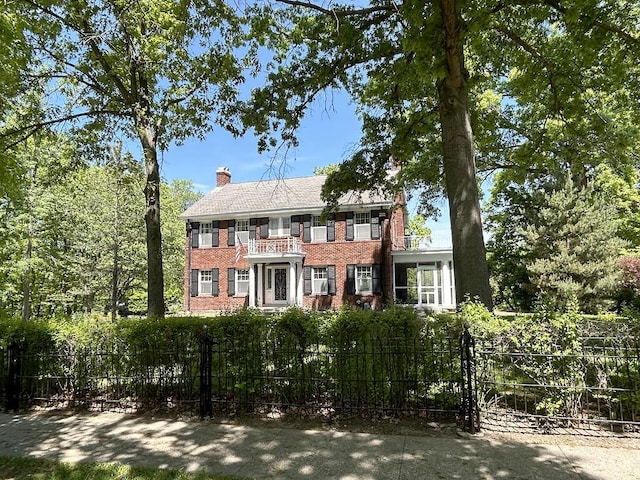 colonial inspired home with a balcony, a fenced front yard, brick siding, and a chimney
