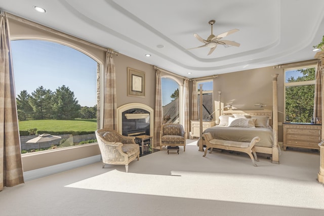 carpeted bedroom featuring recessed lighting, a raised ceiling, multiple windows, a glass covered fireplace, and a ceiling fan