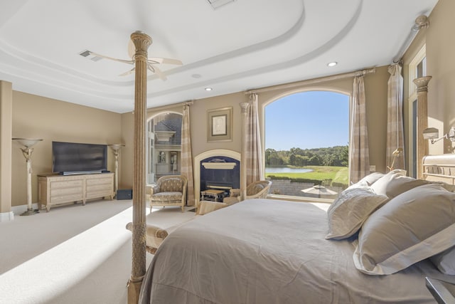 carpeted bedroom with a water view, a fireplace, a tray ceiling, recessed lighting, and a ceiling fan