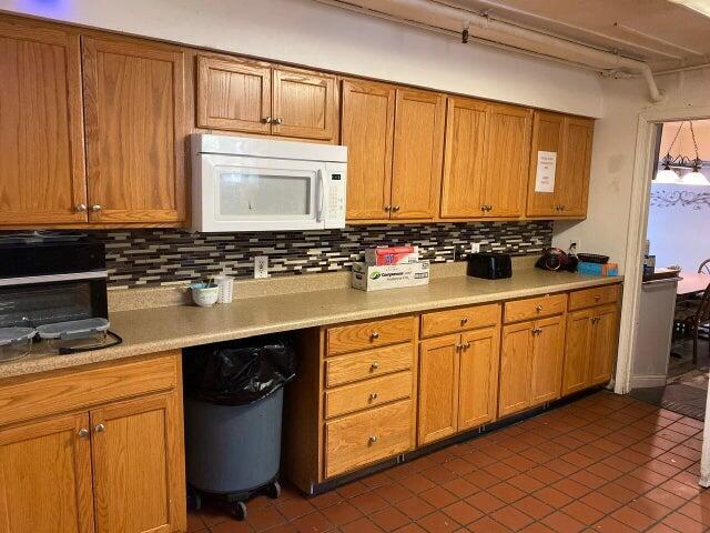kitchen with white microwave, decorative backsplash, brown cabinetry, light countertops, and dark tile patterned flooring