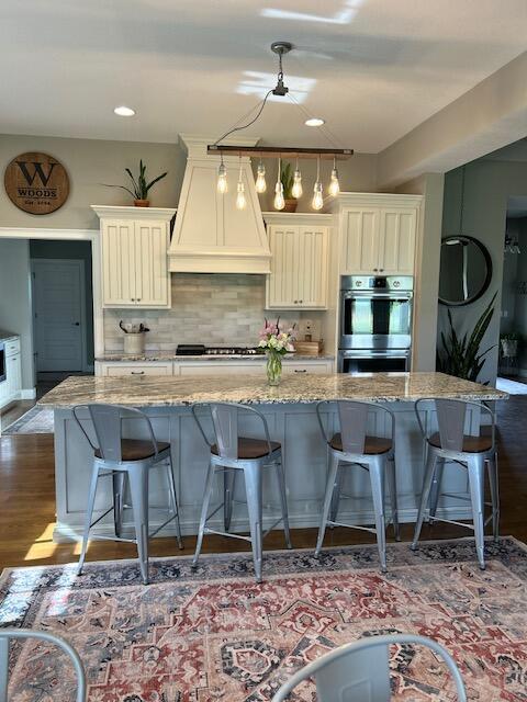 15172 S Henry Ln, Ashland, MO 65010 - kitchen with double oven, custom exhaust hood, light stone countertops, dark wood-type flooring, and a kitchen bar