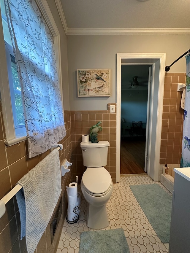 bathroom with tile walls, toilet, a tile shower, ornamental molding, and wainscoting