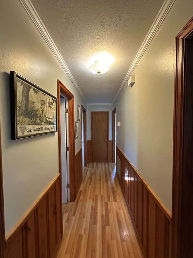 hallway with a textured ceiling, crown molding, wood walls, a wainscoted wall, and light wood-type flooring