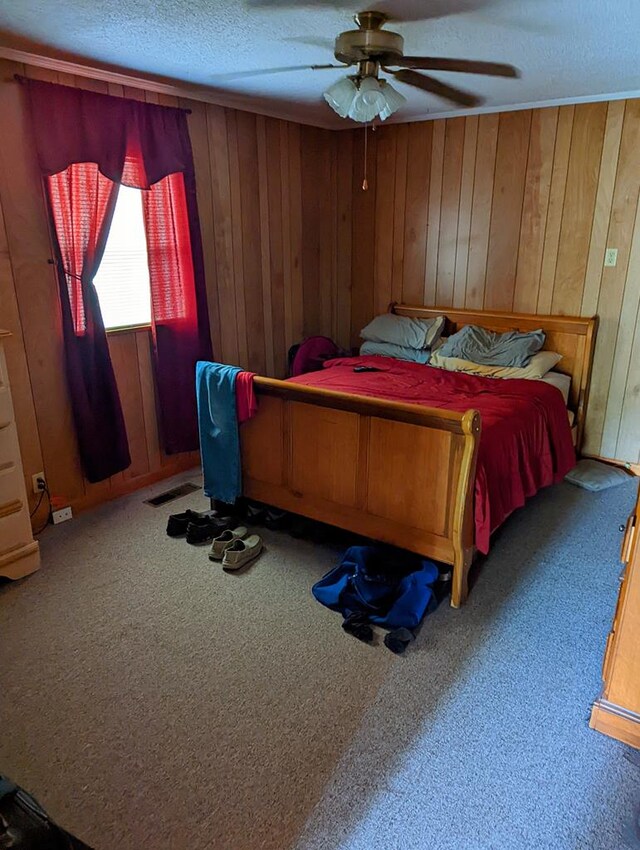 carpeted bedroom with a textured ceiling, wooden walls, a ceiling fan, and visible vents