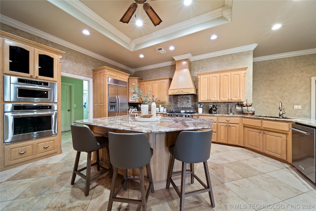 kitchen featuring appliances with stainless steel finishes, an island with sink, custom exhaust hood, ceiling fan, and a tray ceiling