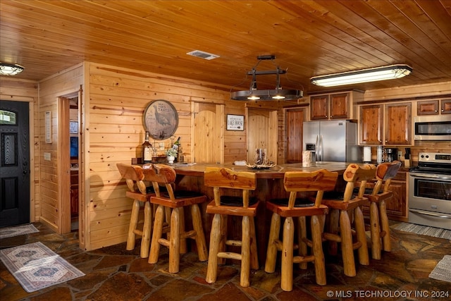 kitchen with stainless steel appliances, wood walls, a kitchen bar, dark tile flooring, and wood ceiling