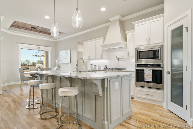 kitchen with appliances with stainless steel finishes, a chandelier, a center island with sink, custom exhaust hood, and light wood-type flooring