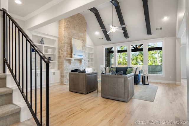 living room featuring built in shelves, light hardwood / wood-style flooring, ceiling fan, a stone fireplace, and beam ceiling