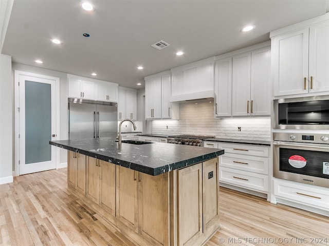 kitchen with sink, tasteful backsplash, light wood-type flooring, a center island with sink, and built in appliances