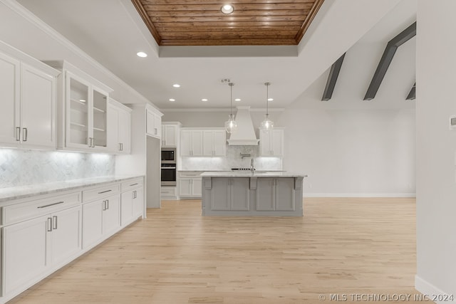 kitchen with an island with sink, custom range hood, glass insert cabinets, light stone countertops, and white cabinetry