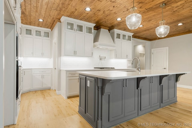 kitchen with wooden ceiling, premium range hood, white cabinets, light wood-type flooring, and an island with sink