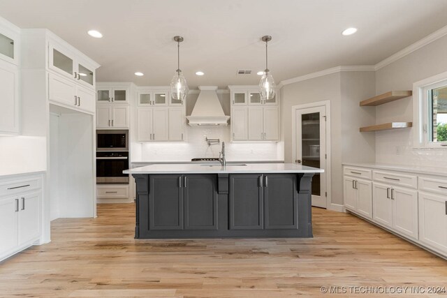 kitchen featuring white cabinets, hanging light fixtures, light hardwood / wood-style flooring, premium range hood, and a kitchen island with sink