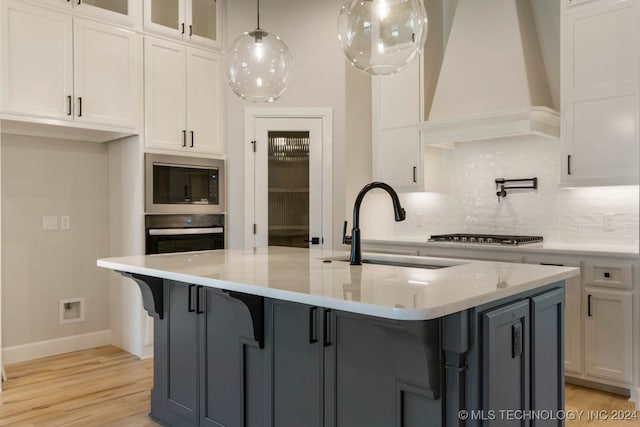 kitchen featuring an island with sink, stainless steel appliances, white cabinetry, and custom range hood