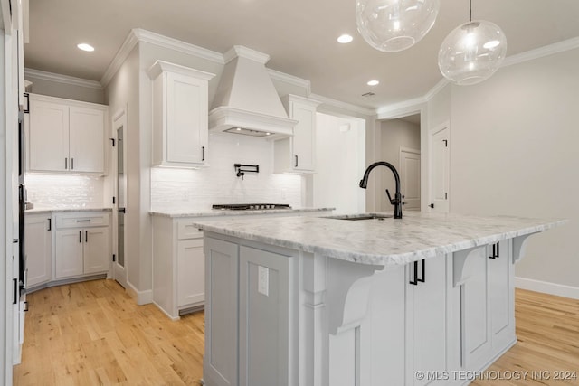 kitchen with an island with sink, premium range hood, white cabinetry, light stone countertops, and light hardwood / wood-style floors