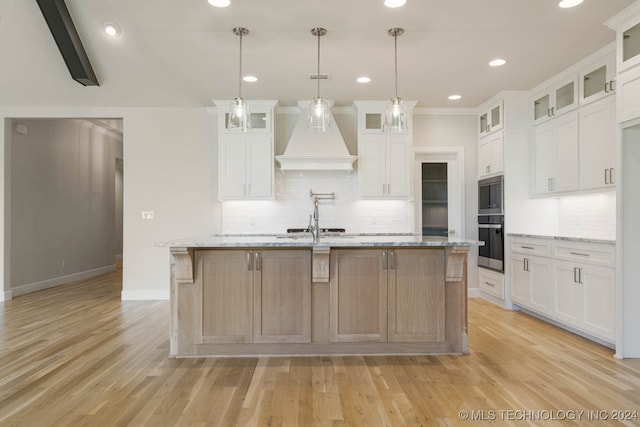 kitchen featuring light wood-type flooring, hanging light fixtures, a center island with sink, white cabinetry, and light stone countertops