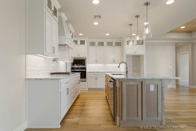 kitchen featuring appliances with stainless steel finishes, white cabinetry, premium range hood, a kitchen island with sink, and sink