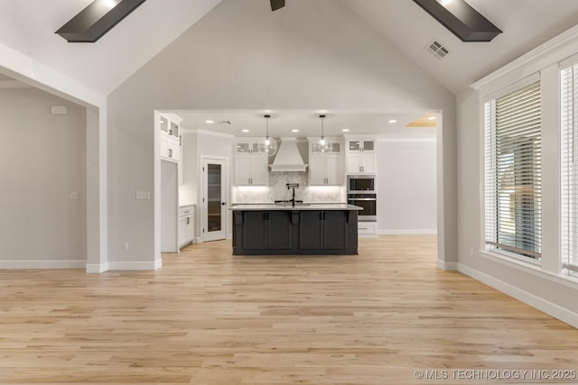 kitchen featuring white cabinets, hanging light fixtures, an island with sink, glass insert cabinets, and custom range hood