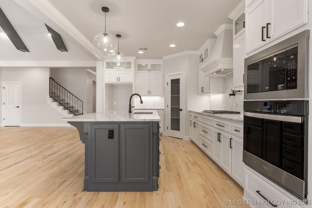 kitchen featuring premium range hood, a center island with sink, a sink, and white cabinetry