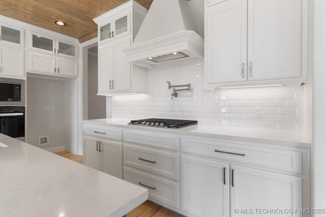 kitchen with stainless steel appliances, decorative backsplash, glass insert cabinets, white cabinetry, and premium range hood