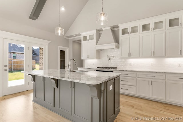 kitchen with white cabinetry, custom range hood, decorative light fixtures, a kitchen island with sink, and light wood-type flooring