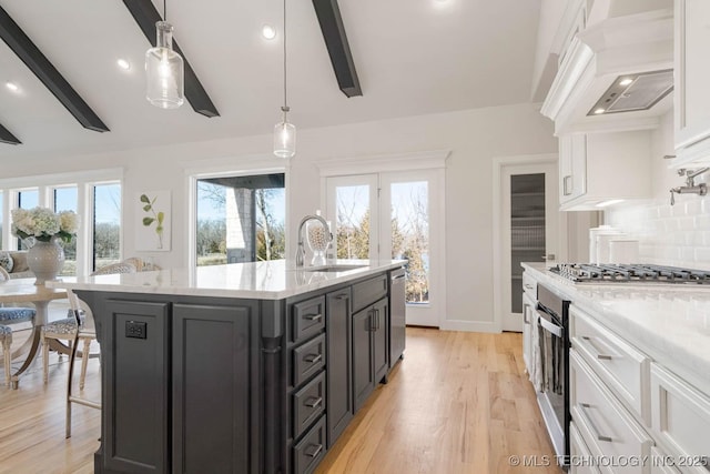 kitchen featuring decorative light fixtures, sink, white cabinets, custom range hood, and a center island with sink