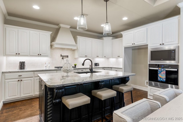 kitchen with a center island with sink, custom exhaust hood, stainless steel appliances, white cabinets, and light stone countertops