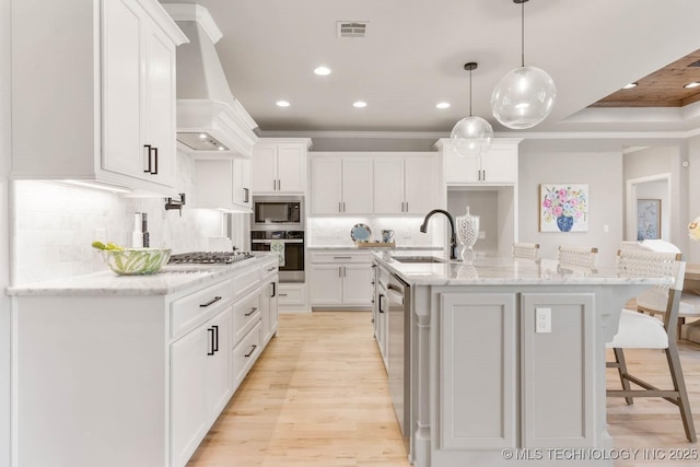 kitchen with custom exhaust hood, stainless steel appliances, white cabinetry, a sink, and an island with sink