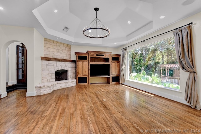 unfurnished living room featuring light wood-type flooring, a tray ceiling, a stone fireplace, and a notable chandelier