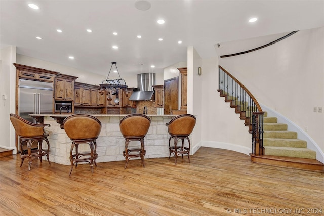 kitchen with wall chimney range hood, light wood-type flooring, light stone counters, kitchen peninsula, and a breakfast bar area