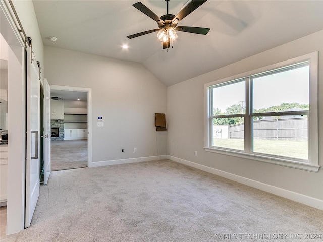 carpeted spare room featuring a stone fireplace, vaulted ceiling, a barn door, and ceiling fan