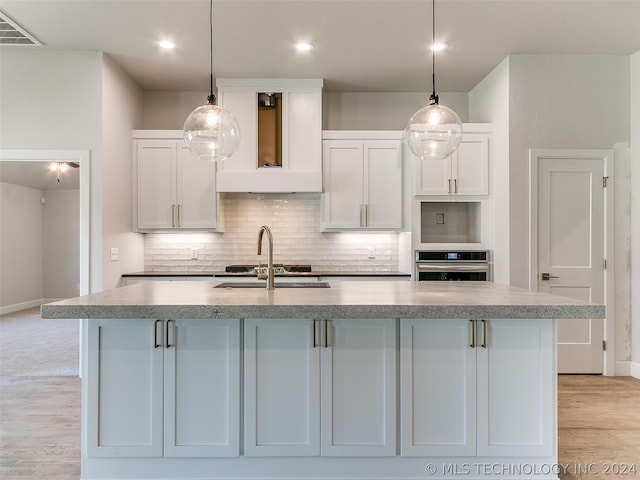 kitchen with stainless steel oven, decorative light fixtures, custom exhaust hood, a center island with sink, and light hardwood / wood-style flooring