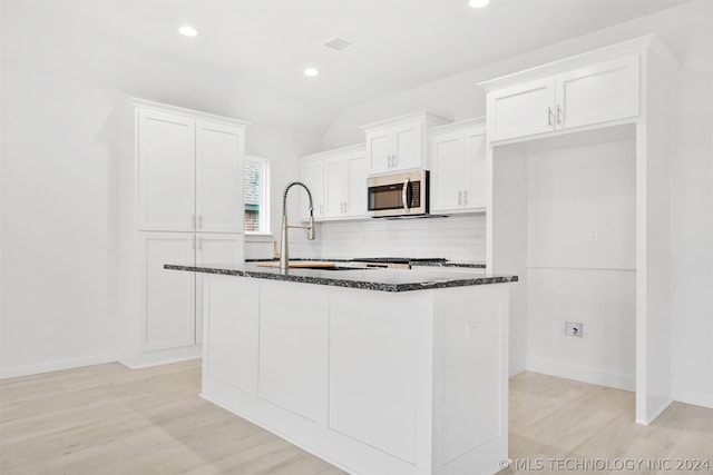 kitchen with light hardwood / wood-style floors, white cabinetry, an island with sink, and dark stone counters