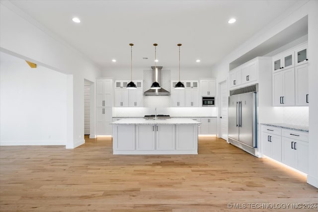 kitchen with hanging light fixtures, stainless steel appliances, wall chimney range hood, a kitchen island with sink, and white cabinets